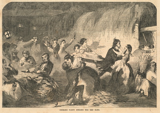 Many people gather to husk corn in a large open barn. Two sets of couples appear to have found red ears of corn and embrace for a kiss.