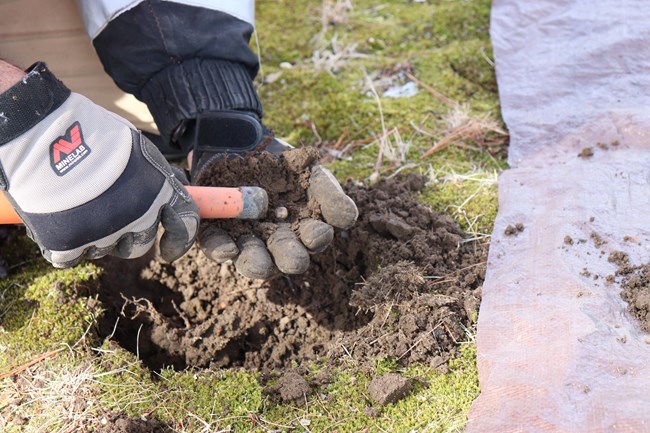 A small white musket ball is held by a hand with a black glove while a small metal detector is pushed toward it.