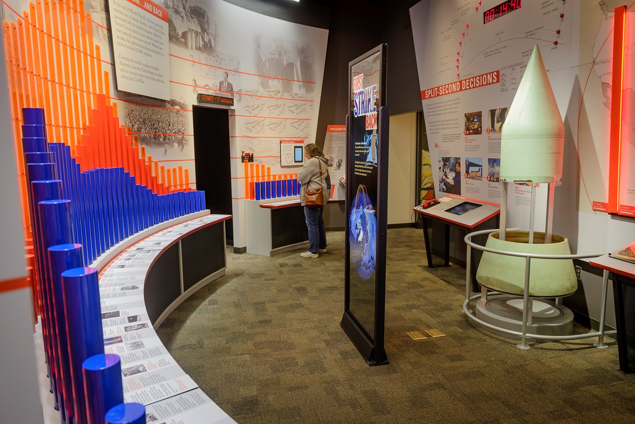 Visitors stand near a curved wall with bright exhibit panels and standing tubes which represent the nuclear arms race