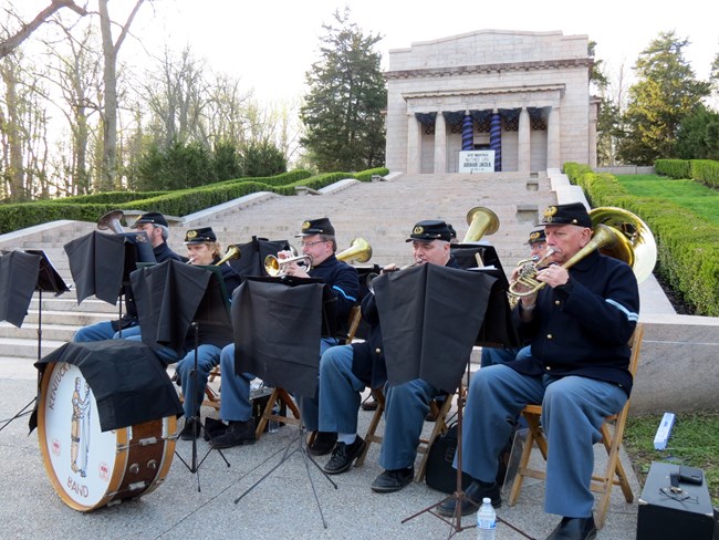 A group of musicians dressed in blue uniforms playing brass instruments with stairs and monument in the background.