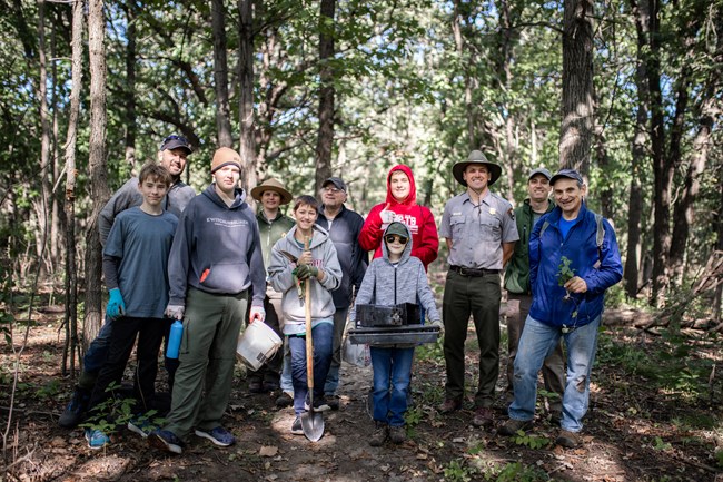 Group of volunteers and park rangers smile while holding planting tools and materials in a wooded work site.