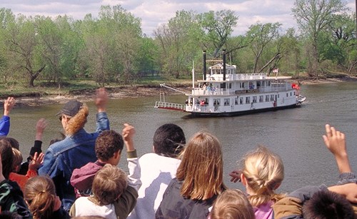 Students on the bank waving at a paddleboat on a river.