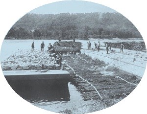 Men pile mats of willow in the river while others prepare to weigh the mat down with rocks.