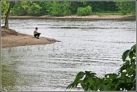 A man sits at the edge of an island in quiet contemplation. The Mississippi and Minnesota Rivers converge here in among tree-lined shores.