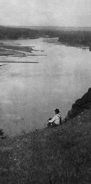 A man sits on a river bluff looking at the river.