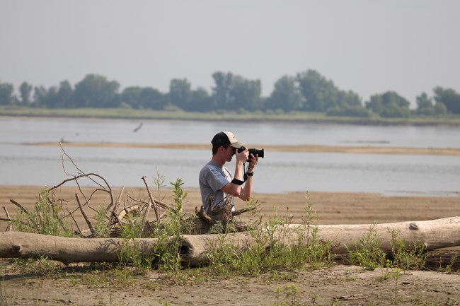 A man holding a black camera kneels down on a sandbar behind a log. The river and its far bank are in the background.