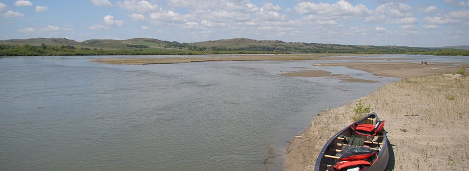 A canoe with red life jackets on a river beach with a river in the background.