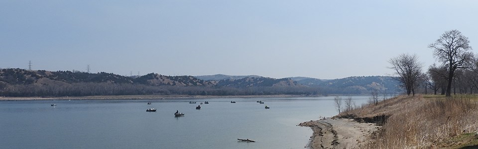 A dozen boaters out on the river fishing near Randall Creek in early spring. The sky is hazy blue. Trees along the river's edge in the background are without leaves. Across the river and beyond the boaters are rolling hills dotted with green cedar trees.