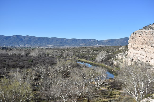 A forested creek winding through a valley sided by a few steep cliffs viewed from one of the cliff faces. A rolling mountain range is visible in the distance.
