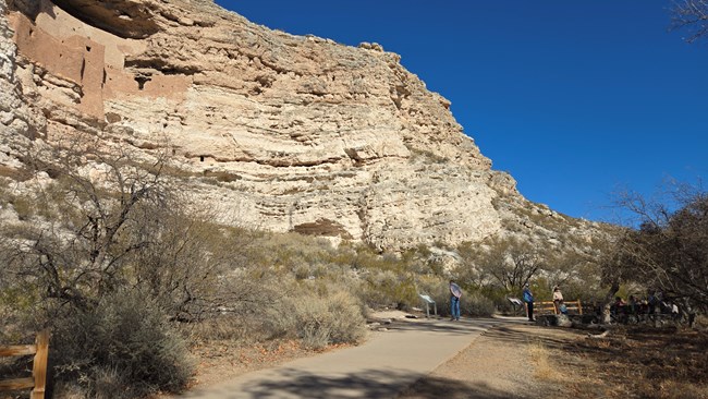 A large, intact cliff dwelling above a wide, paved trail with some signs. A few people are looking at the signs or up at the dwelling.