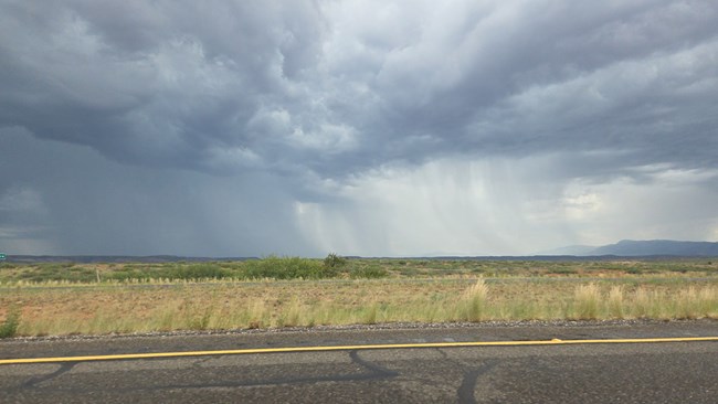 an open, desert landscape with dark clouds in the distance and bands of heavy rain falling