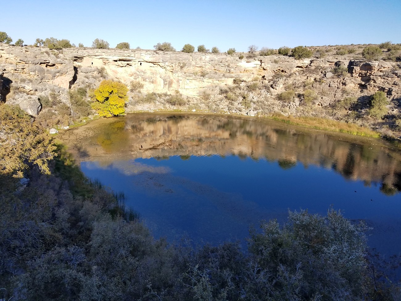 A large sinkhole filled with blue water reflecting the sky. The sinkhole is surrounded by reeds, cattails, shrubs, and trees. Two small stone dwellings are built on a natural ledge on the wall of the far side.
