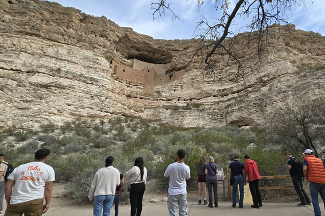 a group of people standing on a trail in front of a large cliff dwelling high above the trail