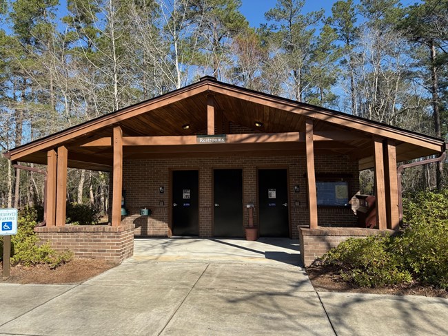 Image of a public restroom facility with three doors, a covered porch, and a sign indicating 'Restrooms'. The building is constructed of brick and wood and is situated in a natural setting with trees and greenery.