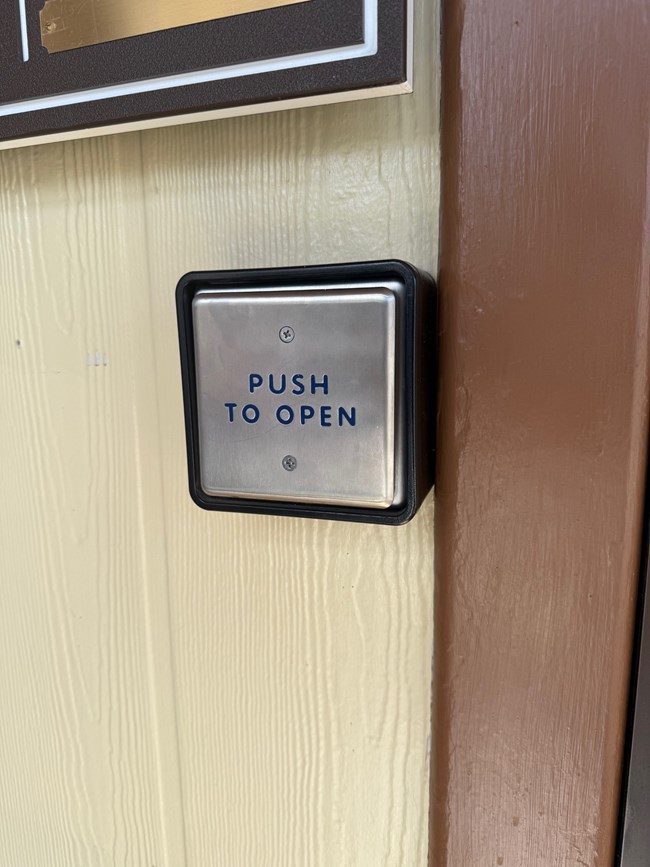 A square metal push plate mounted on a light-colored wall, with the words 'PUSH TO OPEN' in blue lettering. The plate has a black rubber surround and is positioned near a brown door frame.