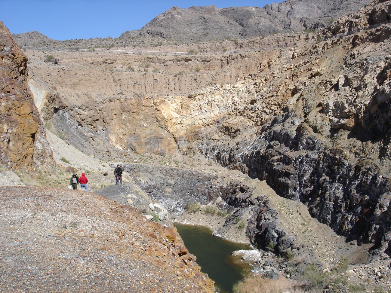 Three people stand on a rock ledge overlooking the deep water-filled pit of the Vulcan Mine