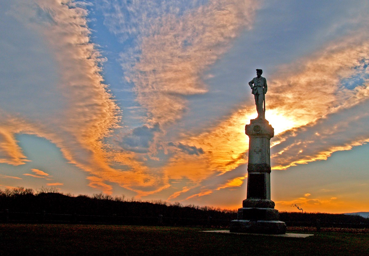 A tall granite column with a statue of a soldier on top is framed by a cloudy sky with streaks of orange. The silhouette of low hills in the background.