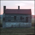 A small two-story building with stone walls on first floor and white on the second story. Rolling farm fields in the background. The setting sun is turning the sky orange and red.