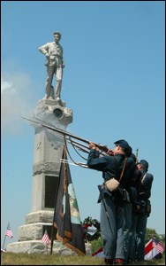 Men in Union army uniforms fire a salute in front of a monument with a stone statue of a Union soldier on top.