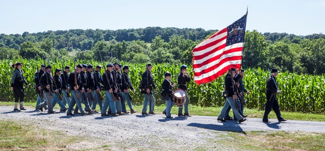 Civil War soldier re-enactors march carrying a large US flag.