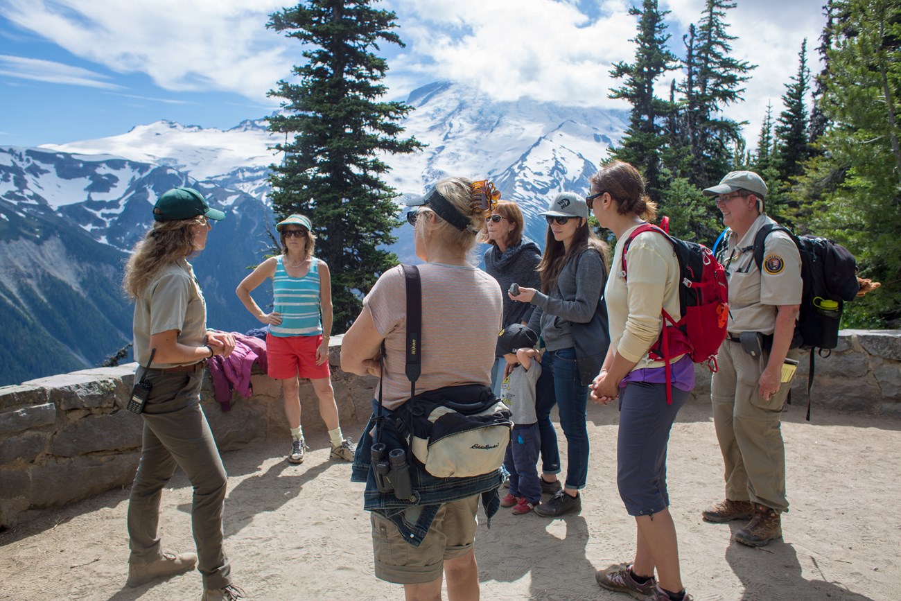 A volunteer ranger talks to a group of visitors at a viewpoint in front of a mountain range.