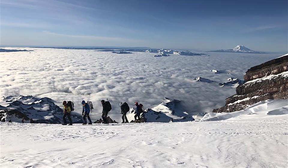 A row of climbers cross a steep snowy slope of a mountain high above a layer of clouds and a distant volcanic peak.