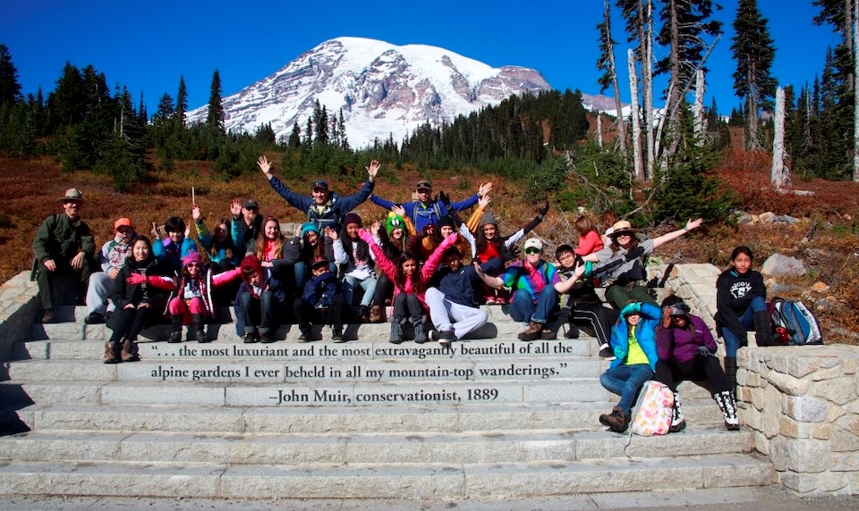 A group of smiling kids, teachers, and rangers sit on stone steps, arms spread wide in welcoming, in front of a view of Mount Rainier.