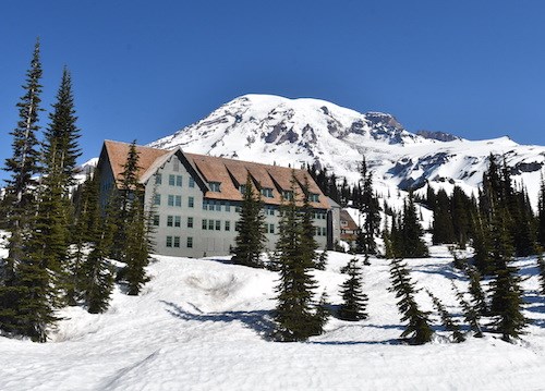 A tall wood building on a snowy slope dotted with fir trees with the peak of a glaciated mountain behind it. .