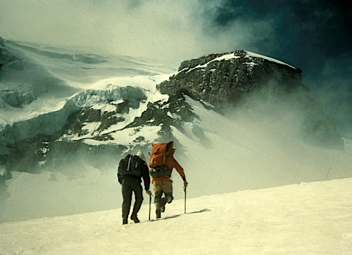 Two climbers ascend a steep snowy slope towards a large rock outcrop partially obscured by dark clouds.