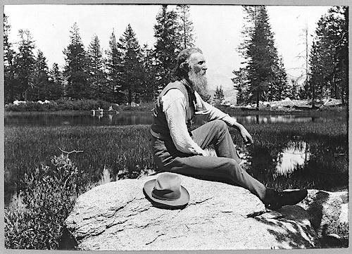 A black and white portrait of a man with a long beard sitting in profile on a large rock next to a pond, his hat on the rock next to him.