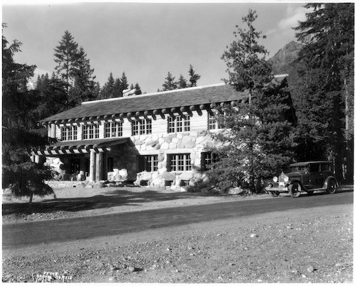 Black and white photo of a two story wood and stone building with a dirt road next to it.