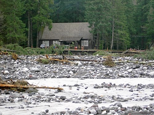 A wide river bed filled with large rocks and fallen trees flows in front of a wood building tucked against a forest.