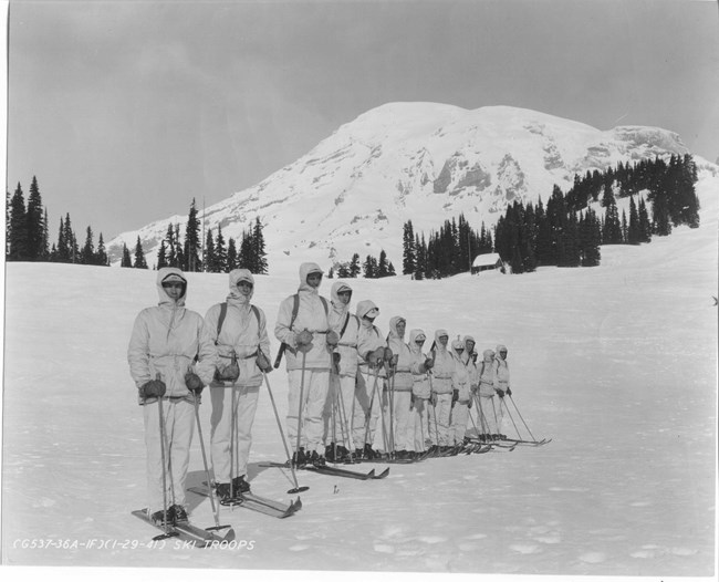 A dozen men on skis in white uniforms with Mount Rainier in the background