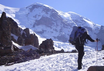A climber with a large backpack and hiking poles walking across snow towards the rocky summit of Mount Rainier.