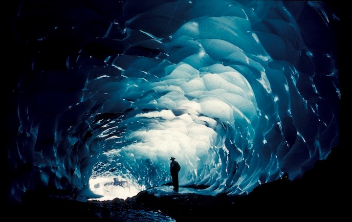 A man stands inside a dark ice cave, silhouetted by light coming from the entrance.
