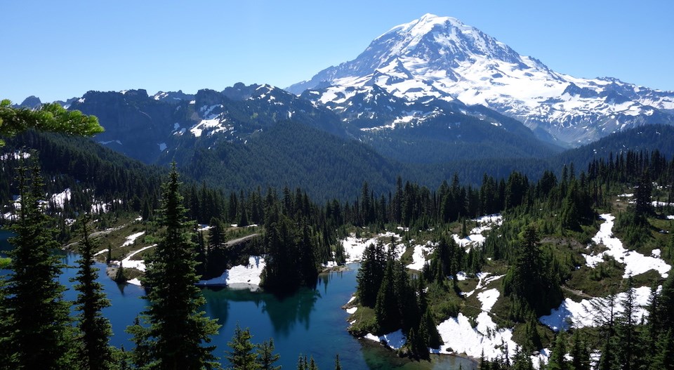 Looking out over a subalpine lake surrounded by patchy snow to the summit of Mount Rainier.