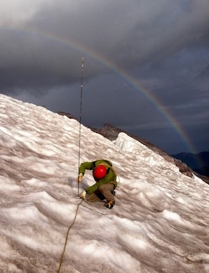 A person kneels on a glacier holding a stake with a rainbow arching overhead.