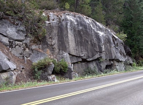 Dark grey rocks, streaked lighter grey in sections, next to a paved road.