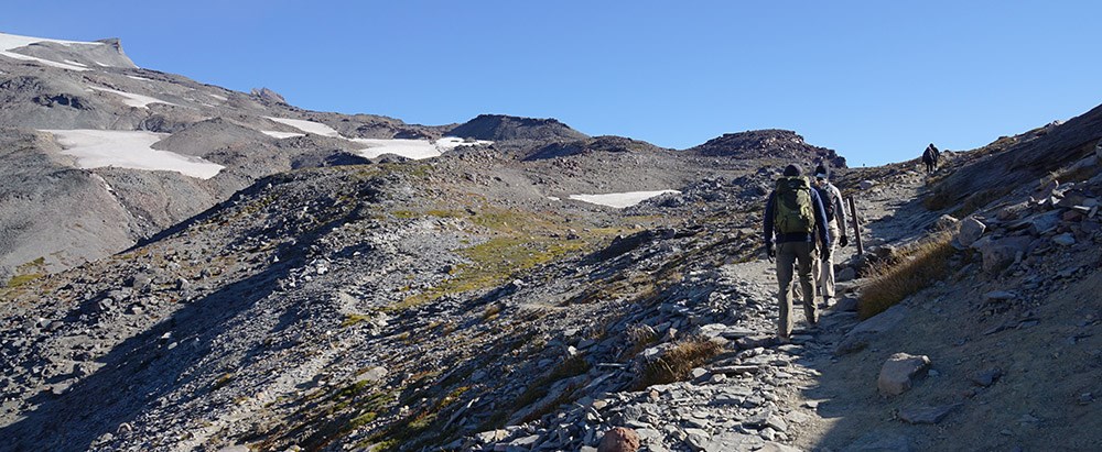 Hikers walk along a rocky trail that follows the ridge of a mountain.