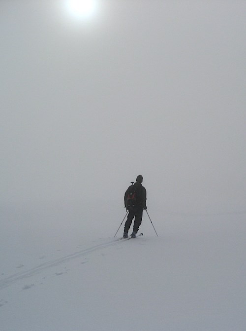A skier pauses on a snowy slope as fog obscures the landscape.