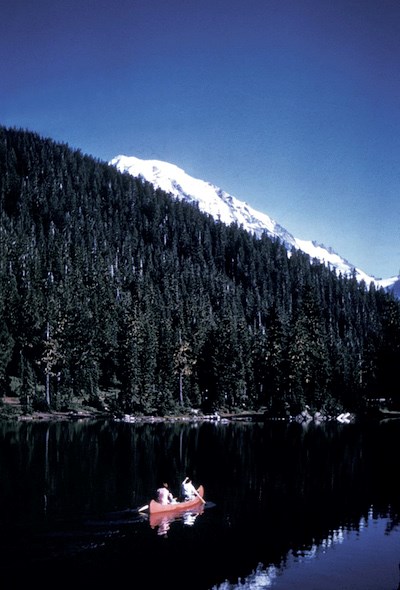 Two people in a red canoe paddle on Mowich Lake, with Mount Rainier barely visible over the edge of the surrounding forested hillside.