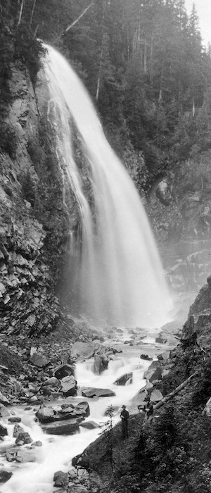 A black and white historic photo of a waterfall with a man standing at its base.