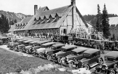 A black and white historic photo of a large wood and stone building with old fashioned cars parked in front.