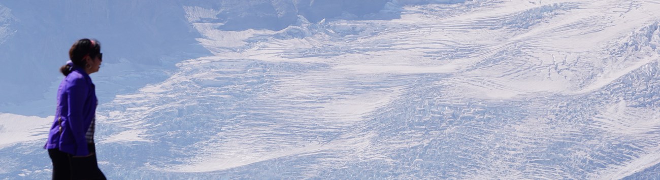 A hiker is in the foreground, slightly out of focus. In the background is sunlit glacial ice.