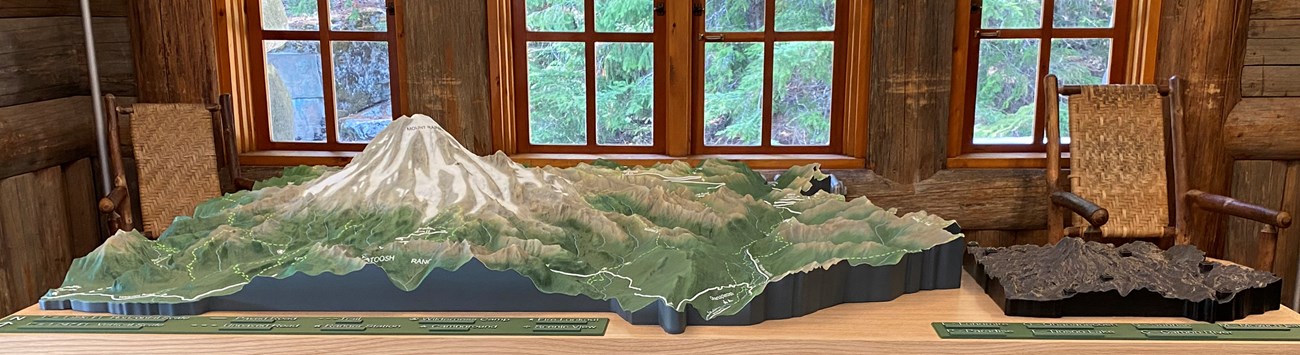 A relief map model of Mount Rainier National Park sits in front of a rustic wall at the Longmire Wilderness Information Center. There are braille panels in the foreground.