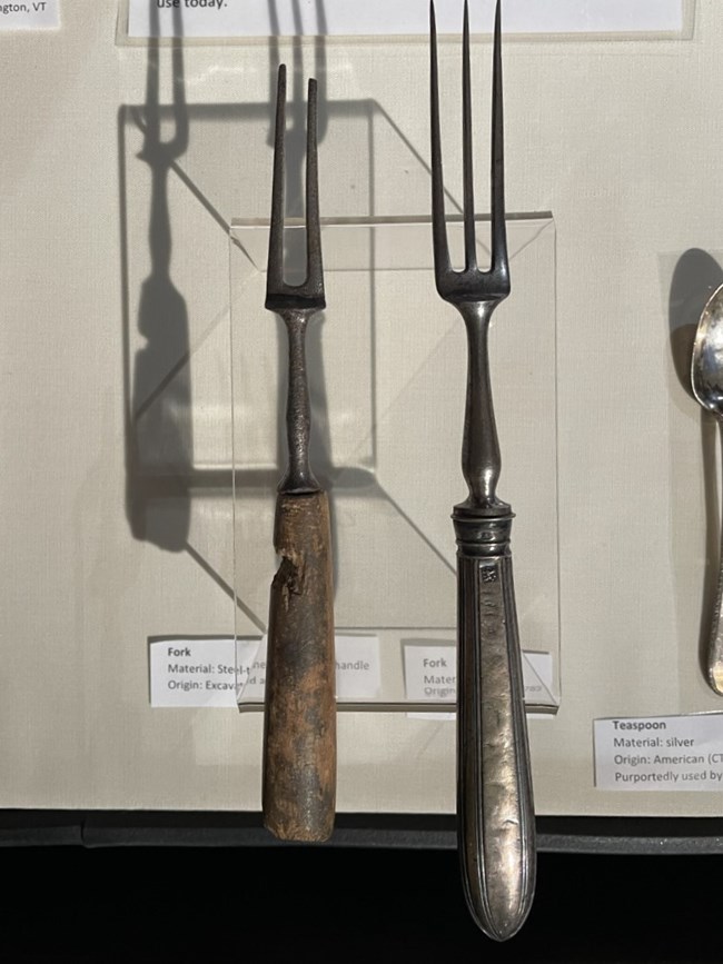 A wooden handled steel fork and a silver fork elevated for display in a museum case.