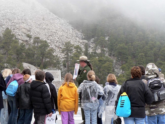 A male park ranger wearing a tan hat and green jacket holds an image and talks to students at Mount Rushmore.  The students are gathered around the ranger.  The talus slope and fog are visible in the background.