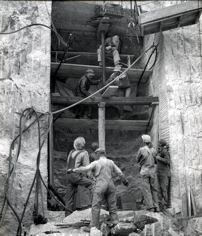 5 workers stand at the bottom of the Hall of Records while 3 more are on scaffolding above.  Construction is in the early stages as the hall is only 6 feet deep.  Wood planks are suspended across the hall providing access to the top of the opening.