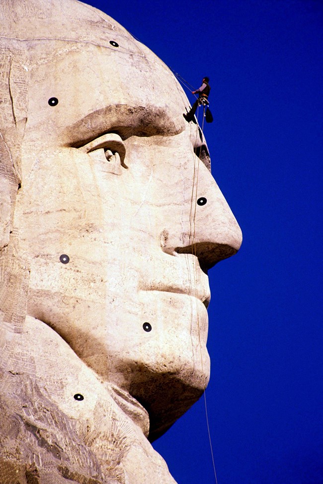 A closeup view of George Washington's carved face looking to the right with a deep blue sky behind.  A worker stands in a harness just above the nose.  Six round black targets are visible to aid in scanning of the sculpture.