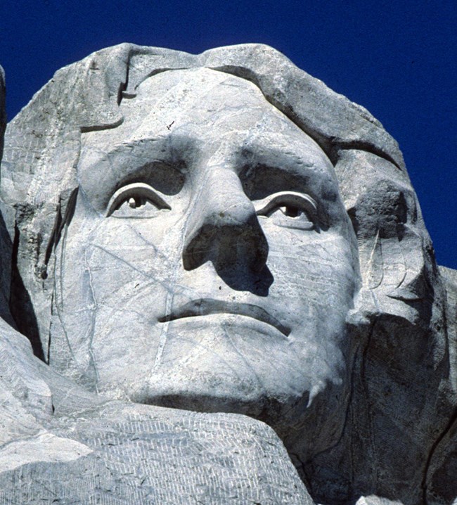 Closeup view of Thomas Jefferson's image carved onto the gray granite on Mount Rushmore.  Numerous veins of lighter colored granite and cracks are visible.  He is looking slightly to the right and up and is depicted with a pleasant expression.